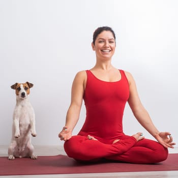 Caucasian woman in red jumpsuit sits in lotus position next to the dog. Yoga with a pet.