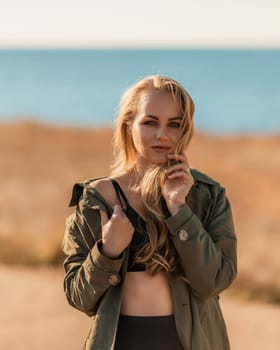 Portrait blonde sea cape. A calm young blonde in an unbuttoned khaki raincoat stands on the seashore, under the raincoat there is a black skirt and top.