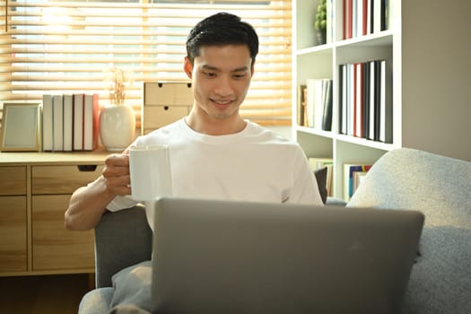 Happy asian male freelancer holding cup of coffee and using laptop on couch. People, technology and lifestyle concept.