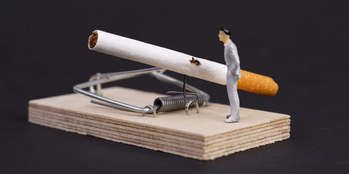 Cigarettes are placed in a mousetrap, next to it is a figure of a man. Trap in the form of nicotine addiction. Dark background.