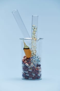 Vertical photo of a graduated laboratory beaker with pills, ampoule and test tubes with therapeutic dragees and translucent capsules, isolated on blue background. World Pharmacist and Health Day.