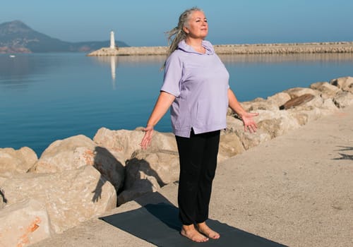 Serious aged woman in with dreadlocks hairstyle standing on a beach against sea with raised arms while practicing pose during sunny day