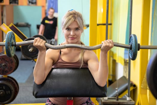 A girl at the gym holds a barbell in front of her. The woman raises the barbell to chest height. A mirror can be seen in the background. In the mirror is the reflection of a blurry person in a black t-shirt