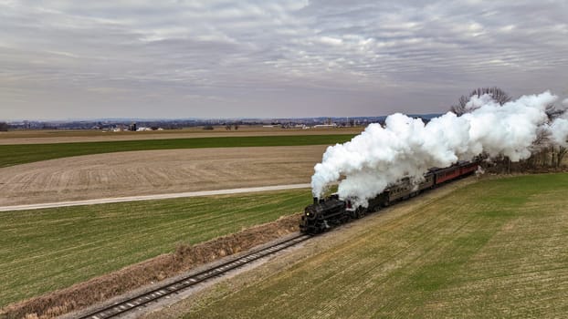 An Aerial View of a Steam Passenger Train Approaching, Traveling Thru Open Farmlands, Blowing Lots of White Smoke, on a Winter Day