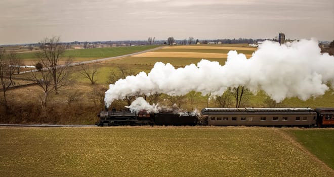A Side Aerial View of a Steam Passenger Train Approaching, Traveling Thru Open Farmlands, Blowing Lots of White Smoke, on a Winter Day