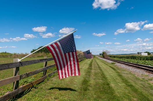 A Restored Antique Steam Passenger Train Approaches a Fence With Gently Waving American Flags on It On a Sunny Summer Day