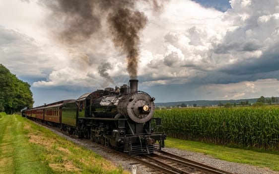 A Frontal View of an Antique Passenger Train Approaching Blowing Black Smoke on a Summer Day With Storm Clouds Approaching