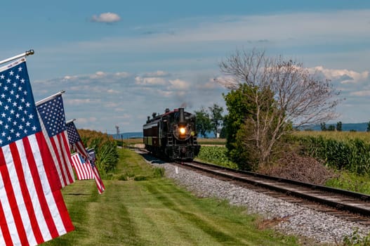 A Restored Antique Steam Passenger Train Approaches a Fence With Gently Waving American Flags on It On a Sunny Summer Day