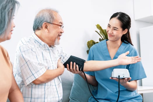 An elderly man having a blood pressure check by his personal caregiver with his wife sitting next to him in their home.