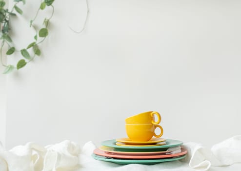 Empty yellow cups and brightly colored plates on a white table covered with a linen tablecloth, a flower pot on the wall in the background. Preparation for serving