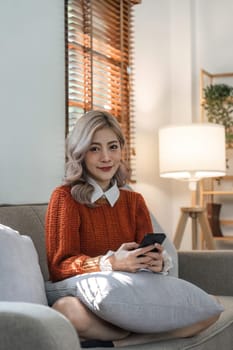 Happy young asian woman on mobile phone while sitting a couch in living room at home, Shopping online via website.