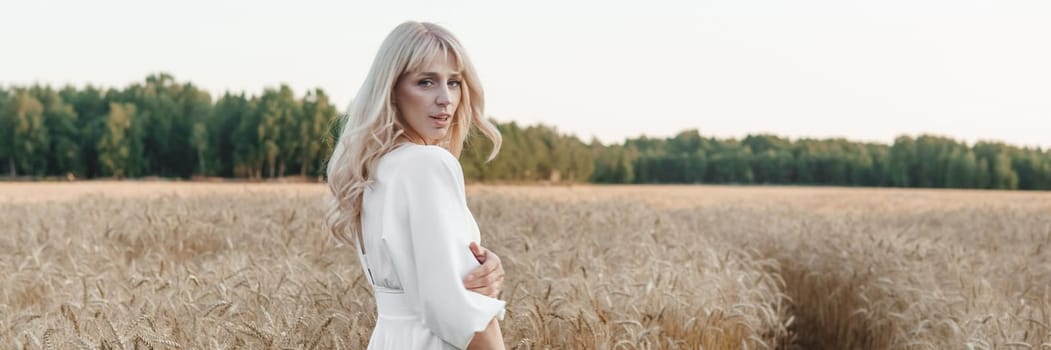 A blonde woman in a long white dress walks in a wheat field. The concept of a wedding and walking in nature.