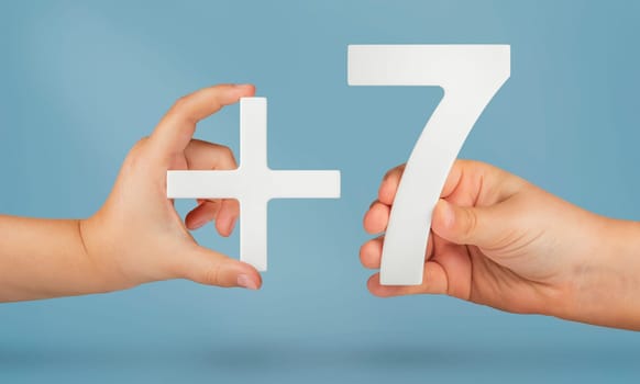 The number seven and the plus symbol in the hands of a child on a blue background. White number 7 with a plus close-up. The concept of addition or sum of the number seven