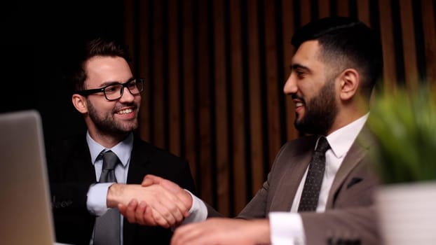 Two young men smile and shake hands as they meet at the table in the company office