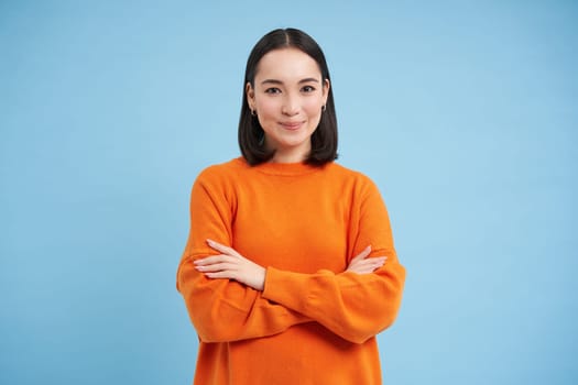 Young asian woman looks with confidence, cross arms on chest and smiles at camera, stands in orange sweatshirt, blue background.