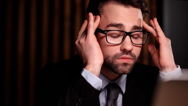 Tired exhausted man, office worker, manager or freelancer, sitting at his desk, tired of working in a laptop, overworked, having a headache, closed his eyes, needs rest and break