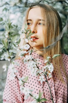 Blonde girl on a spring walk in the garden with cherry blossoms. Female portrait, close-up. A girl in a pink polka dot dress