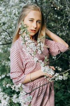 Blonde girl on a spring walk in the garden with cherry blossoms. Female portrait, close-up. A girl in a pink polka dot dress