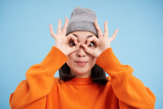 Close up portrait of funny Chinese girl, looks through hand glasses with surprised face expression, standing over blue background.