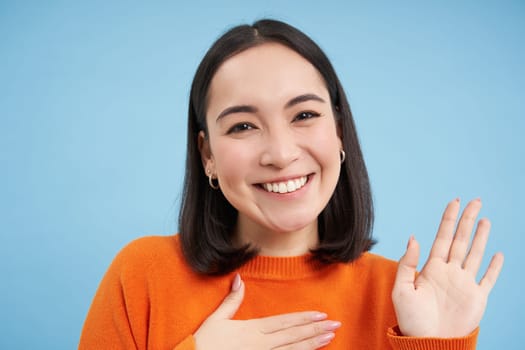 Close up of friendly smiling japanese woman, raises one arm, introduce herself, greets, stands over blue background.