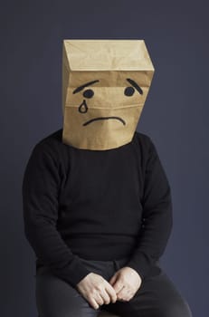 A sad man in a black turtleneck with a bag on his head, with a drawn crying emoticon, sits on a chair. Emotions and gestures. Vertical frame.