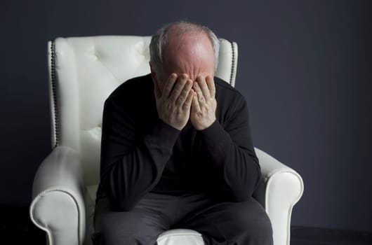 A man in a black turtleneck is sitting in a white chair, rubbing his face with his hands, holding his head. Depression, headache. Medical concept.