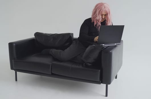 Portrait of a man in a pink wig, sitting on a sofa, typing on a laptop. White background.