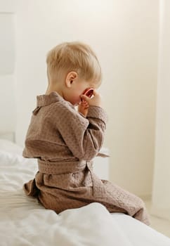 A boy in a brown bathrobe sits on the bed, plays - he closed his eyes with candied oranges. Side view. Vertical frame.