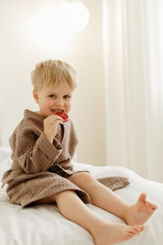 Portrait of a boy in a brown bathrobe who sits on a white bed and eats candied oranges. Vertical frame.