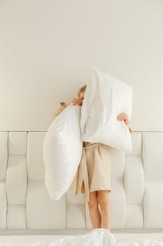 A boy in a bathrobe with pillows in his hands is standing on the bed.