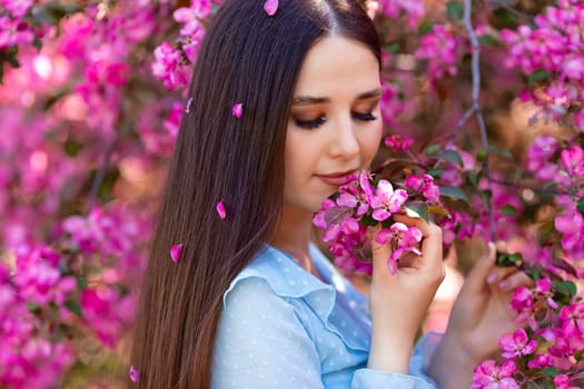 A beautiful brunette girl with pink petals in her long hair stands near a pink blooming apple tree, on a sunny day.