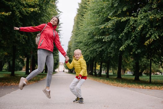 Mother and son jumping in the autumn park. Fall Season. Happy young beautiful mother and her son have a walk in the park