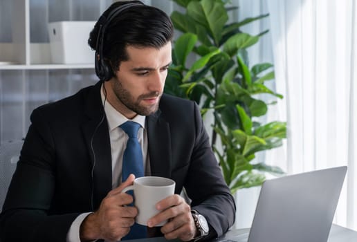 Manager of call center operator office sitting on his desk with his coffee while working on laptop. Office worker wearing headset and black suit working on customer support or telemarketing. fervent