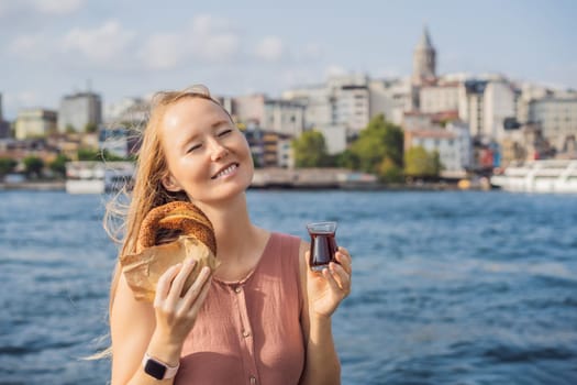 Woman in Istanbul having breakfast with Simit and a glass of Turkish tea. Glass of Turkish tea and bagel Simit against golden horn bay and the Galata Tower in Istanbul, Turkey. Turkiye.