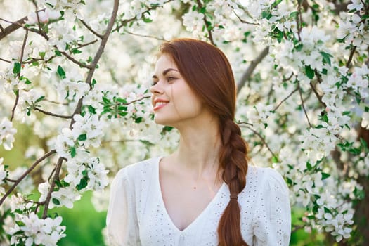 beautiful red-haired woman in a white dress smiling stands near a flowering tree. High quality photo