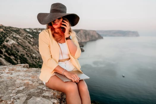 Successful business woman in yellow hat working on laptop by the sea. Pretty lady typing on computer at summer day outdoors. Freelance, travel and holidays concept.