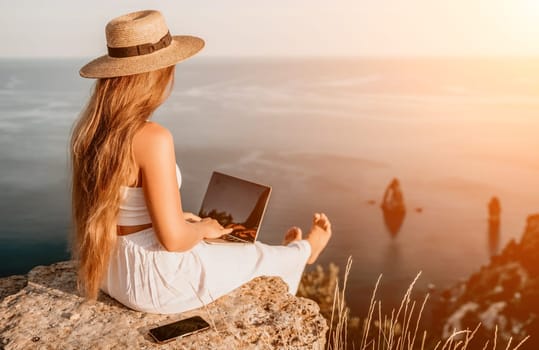 Successful business woman in yellow hat working on laptop by the sea. Pretty lady typing on computer at summer day outdoors. Freelance, travel and holidays concept.