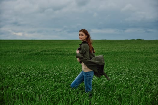 a red-haired woman stands in a green field in rainy, cold weather, holding a raincoat in the wind. High quality photo