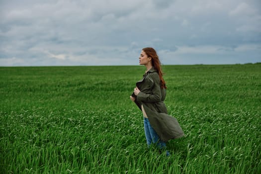 a red-haired woman stands in a green field in rainy, cold weather, holding a raincoat in the wind. High quality photo