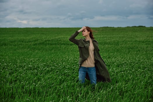 a woman in a dark coat stands in a green field against a cloudy sky holding her hair in the wind. High quality photo