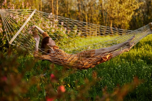 a beautiful woman is resting in nature lying in a mesh hammock in a long orange dress looking to the side, arms outstretched. Horizontal photo on the theme of recreation. High quality photo