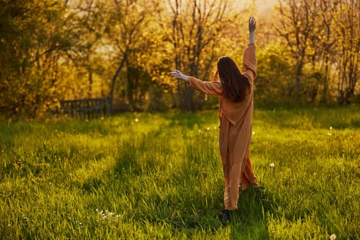a slender woman with long hair stands in a field with her back to the camera, illuminated by the rays of the setting sun and joyfully raises her hands up. Horizontal photography on the theme of unity with nature. High quality photo