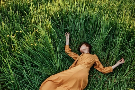 a sweet, calm woman in an orange dress lies in a green field with her arms outstretched and her eyes closed, enjoying the silence and peace. Horizontal photo taken from above. High quality photo