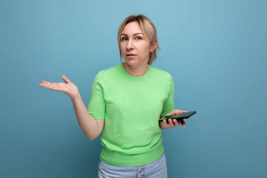 doubtful attractive blond girl holding a smartphone in her hand on a blue background with copy space.