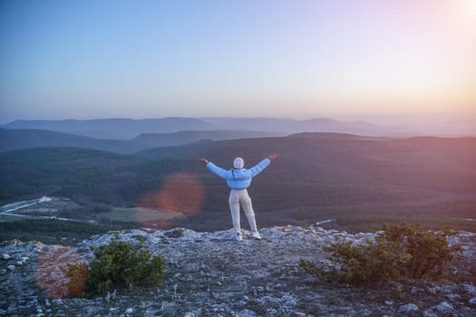 Woman hiker open arms on top of sunrise mountain. The girl salutes the sun, wearing a blue jacket, white hat and white jeans. Conceptual design