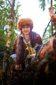 Girl in a leather jacket, a big red fox fur hat in the forest in autumn. A female model poses as a fabulous royal huntress on nature hunt at a photo shoot