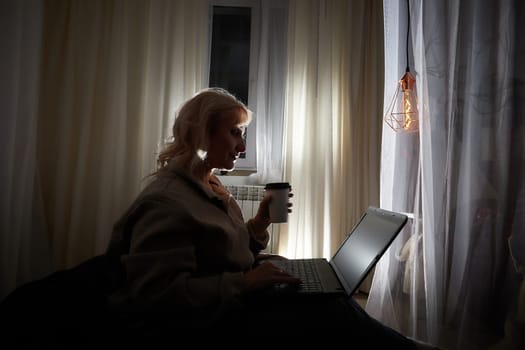 Silhouette and shadow of Adult mature woman of 40-60 years with laptop, notebook, computer in warm sweater in dark calm cozy evening atmosphere room. Interior with curtains and small linght