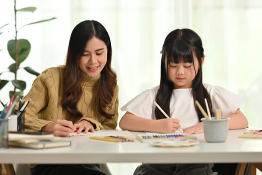 Young adult asian mother and little cute daughter are sitting at table and doing homework together. Learning, education concept.