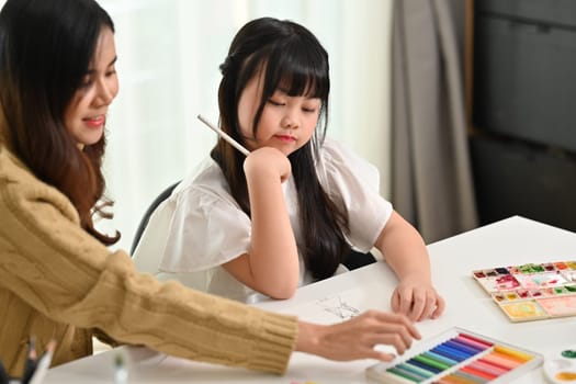 Happy adorable little girl sitting at table doing homework, coloring pages with mother or tutor. Learning, education concept.