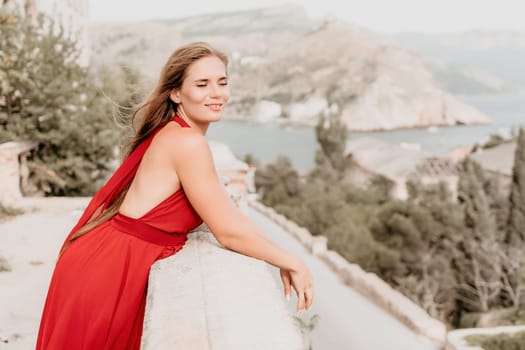 Side view a Young beautiful sensual woman in a red long dress posing on a volcanic rock high above the sea during sunset. Girl on the nature on overcast sky background. Fashion photo